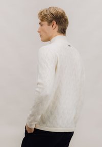 Hvit kabelstrikket genser med høy hals og ribbestrikkede mansjetter, brukt av en modell i profil mot en nøytral bakgrunn.