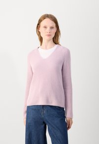 Pulover tricotat de culoare roz pal, cu decolteu în V, textură cu bumb de lână, mâneci lungi și o croială lejeră, purtat peste un tricou alb.