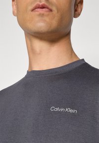 Šedé triko s kruhovým výstřihem, na kterém je na levé straně hrudi bílé vyšívané logo "Calvin Klein", vyrobené z měkké texturované látky.