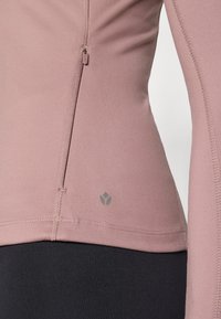 Long-sleeve athletic top in muted pink fabric, featuring a side zipper, grey logo detail, and flatlock seams for smooth texture.