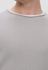 Gros plan d'une personne portant un pull en maille texturée gris clair par-dessus un t-shirt blanc, montrant le cou et le haut de la poitrine.