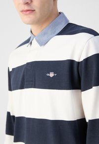 Tamsiai mėlynas ir baltas dryžuotas polo marškinėlis su šviesiai mėlyna apykakle, ant krūtinės mažas logotipas ir tekstūruota medžiaga.
