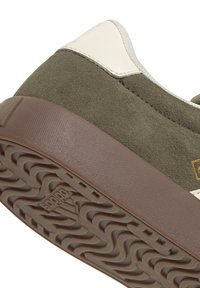 Gros plan sur une basket en daim marron avec une semelle marron texturée et un talon en cuir blanc, montrant le logo Adidas sur la semelle.
