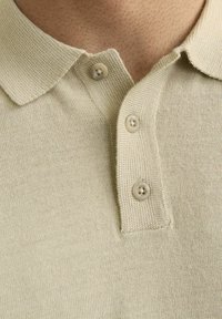 Polo beige avec un tricot texturé, présentant un col classique et deux boutons de couleur claire. Gros plan de la zone du col.