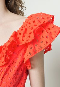 Top vermelho brilhante, com ombros à mostra e mangas curtas com babados. O tecido apresenta recortes circulares, adicionando textura e uma aparência leve e arejada.