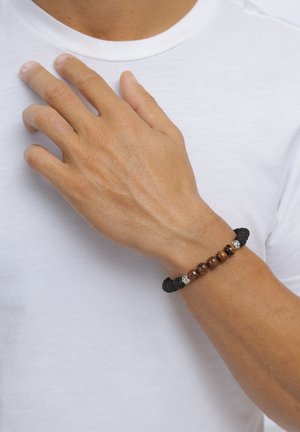 Linke Hand ruht auf der Brust und trägt ein Armband mit braunen, schwarzen und silbernen Perlen am Handgelenk über einem weißen Hemd.