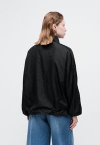 Person med lyst brunt bølget hår iført guld hoops øreringe, en løs sort jakke og vide blå denim bukser, vendt væk mod ensfarvet baggrund.