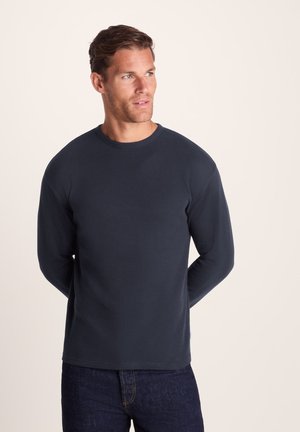Man with short hair wearing a dark long-sleeve textured sweater and dark jeans, looking to his side against a neutral background.