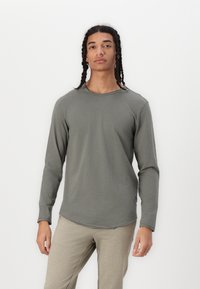 Langärmliges Shirt in olivgrün, aus weichem Stoff mit rundem Ausschnitt und raw hem Detail, getragen mit hellbeigen Hosen.