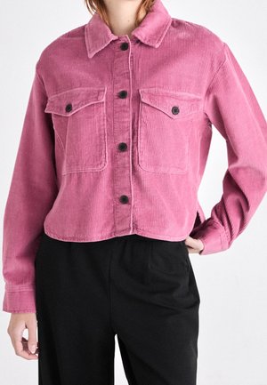 Kobieta ubrana w różową kurtkę na guziki z sztruksu z kieszeniami na piersi oraz czarne spodnie na jednolitym białym tle.