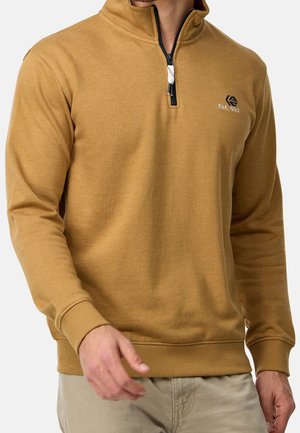 Hombre con sudadera de media cremallera color camello con el logo "Est. 1993", combinado con pantalones beige, posando frente a un fondo blanco.