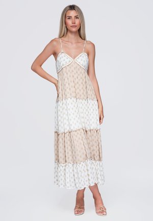 Femme debout portant une robe maxi beige et blanche à étages sans manches, avec bretelles spaghetti et talons ouverts, sur un fond uni.