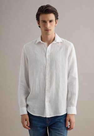 Junger Mann mit dunklen Haaren, der ein weißes, langärmeliges Hemd mit Knöpfen und blaue Jeans trägt, vor einem schlichten beigen Hintergrund stehend.