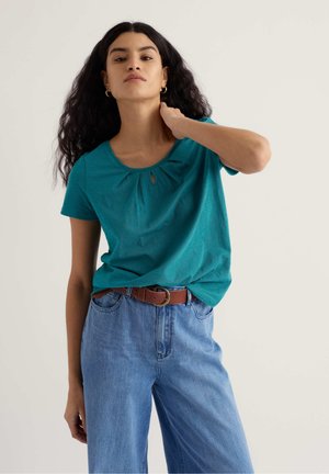 Noor naine pikkade lokkis juustega, seljas türkiissinine lühikeste varrukatega pluus ja sinised teksapüksid pruuni vöös, puudutades kaela ühe käega tavalise tausta ees.