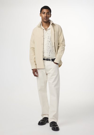 Beige knäppskjorta över en vit mönstrad skjorta, kombinerad med krämfärgade byxor och svarta skor, vilket skapar en lagrad, neutral outfit.