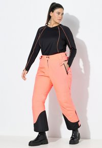 Zwarte lange mouw top met oranje stiksel, gecombineerd met oranje en zwarte geïsoleerde broek. Heeft zakken en een strakke uitstraling. Zwarte laarzen.