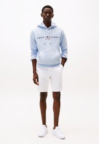 Hanorac albastru deschis cu glugă și logo contrastant, asortat cu pantaloni scurți albi și adidași albi. Material neted, croială casual, design minimalist.