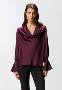 Ljubičasta satenasta bluza s okruglim izrezom, dugim rukavima i detaljem na rukavima, uparen s uskim crnim hlačama.