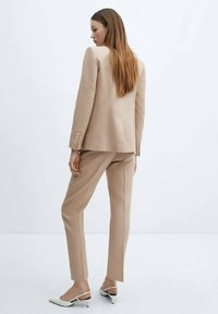 Completo beige chiaro con giacca su misura e pantaloni slim-fit, realizzato in tessuto testurizzato; presenta dettagli con bottoni e tacchi con cinturini alla caviglia.