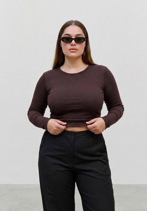 Femme portant des lunettes de soleil foncées, un crop top marron à manches longues et un pantalon noir, debout devant un fond clair uni.