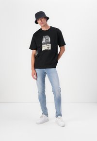 Camiseta de algodón negra con una impresión fotográfica de un vehículo y el texto "TODO TERRENO", combinada con unos jeans de mezclilla azul claro y zapatillas blancas.