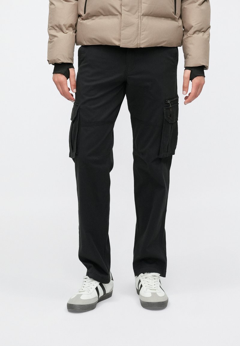 Μαύρο παντελόνι cargo με πολλαπλές τσέπες, ίσια γραμμή και ανθεκτικό ύφασμα. Συνδυασμένο με λευκά αθλητικά παπούτσια με μαύρες λεπτομέρειες.