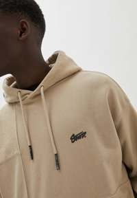 Sweat à capuche beige avec une capuche texturée, une poche avant et des cordons de serrage. Présente un logo noir sur la poitrine. Tissu lisse avec une texture contrastante.