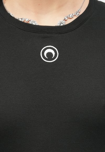 Zbliżenie czarnego prążkowanego topu z białym, okrągłym haftowanym symbolem na wysokości klatki piersiowej oraz widoczną częścią srebrnego naszyjnika z kamieniami przy dekolcie.