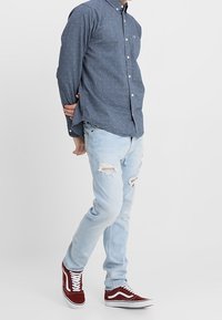 Camisa azul de botones con pequeños lunares blancos, combinada con jeans claros rasgados y zapatillas de ante rojas con cordones blancos.