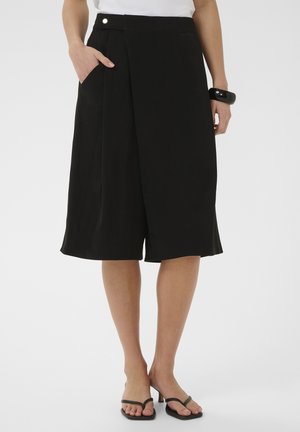 Modell fekete térdig érő, széles szárú sortot visel zsebbel, fehér felsővel, fekete karkötővel és fekete, nyitott orrú magassarkúval, fehér háttér előtt állva.