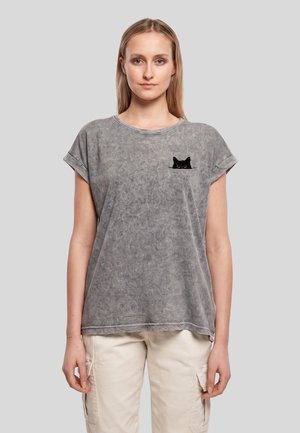 Femme portant un t-shirt gris délavé avec un petit motif de chat noir sur la poitrine gauche et un pantalon cargo beige clair sur un fond uni.