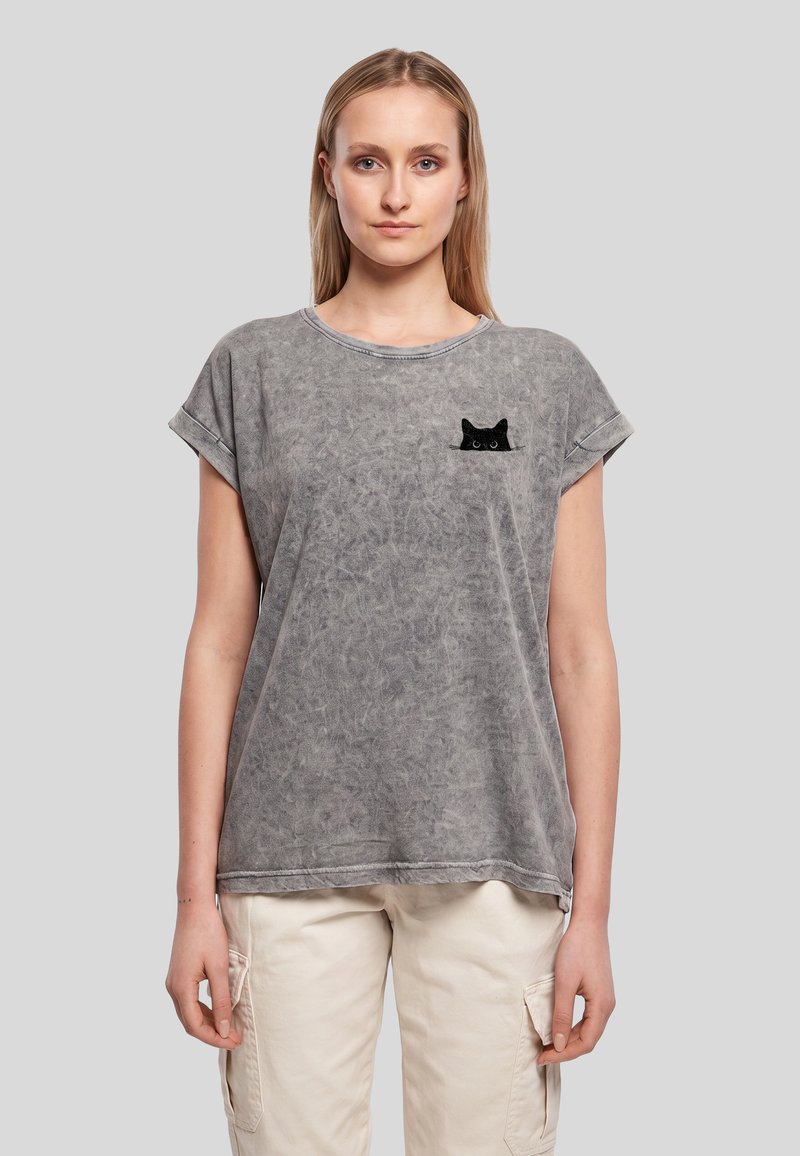Femme portant un t-shirt gris délavé avec un petit motif de chat noir sur la poitrine gauche et un pantalon cargo beige clair sur un fond uni.