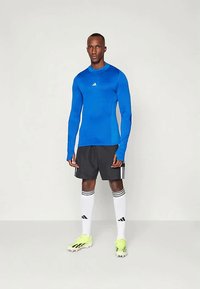 Hosszúujjú kék sportpóló, fekete rövidnadrág, fehér zoknik fekete csíkokkal, és élénk sárga focicipők. Adidas logo a pólón.