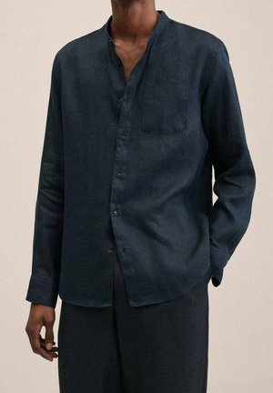 Homme portant une chemise bleu foncé à manches longues avec une poche poitrine, debout avec une main dans sa poche devant un fond uni.