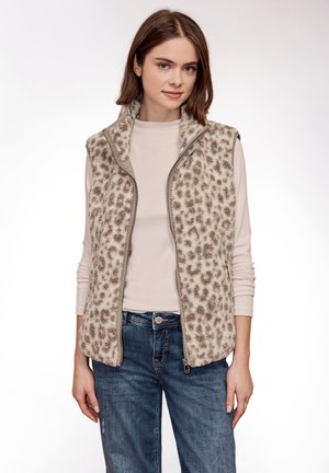 Jeune femme portant une chemise beige à manches longues, un gilet en polaire imprimé léopard et un jean bleu, debout devant un fond blanc uni.