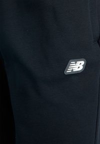 Čierne tepláky vyrobené z jemného materiálu, s malou šedou nášivkou loga New Balance na boku, s hladkou textúrou a jednoduchým dizajnom.