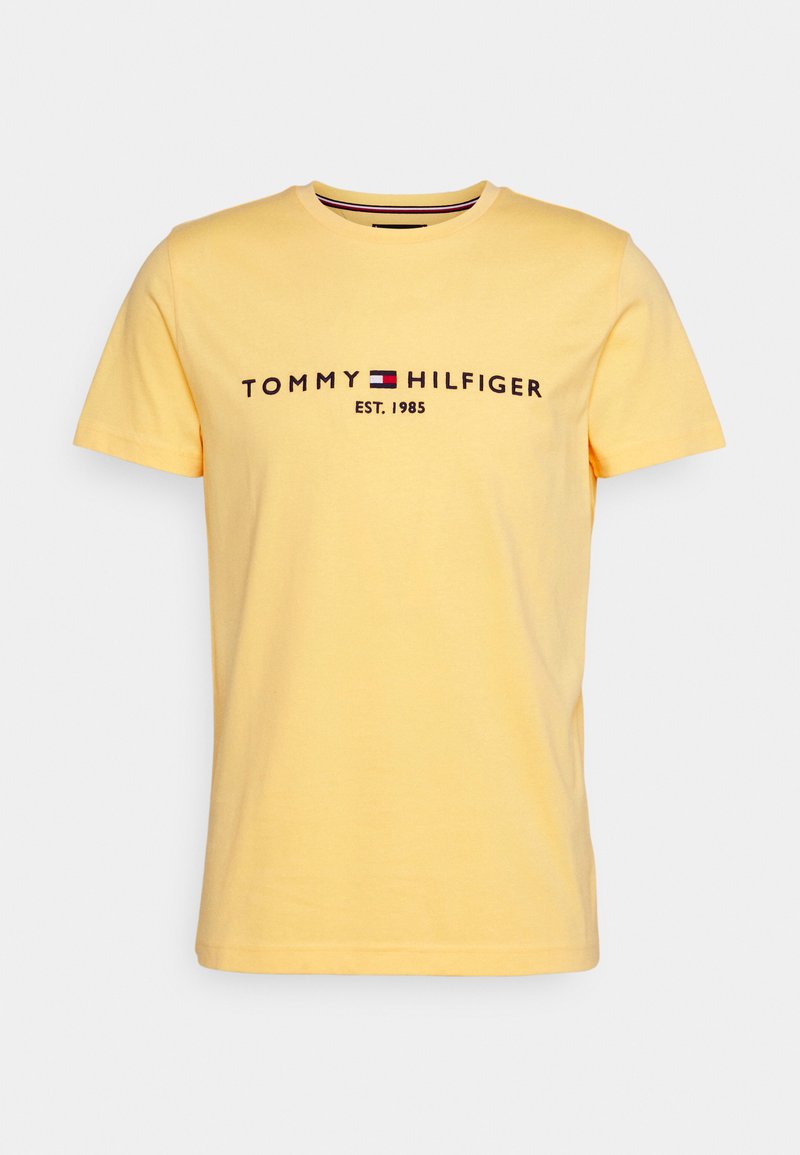 T-shirt de algodão amarelo com mangas curtas. Apresenta o texto preto "TOMMY HILFIGER EST. 1985" e um pequeno detalhe de logotipo nas cores vermelho, branco e azul.