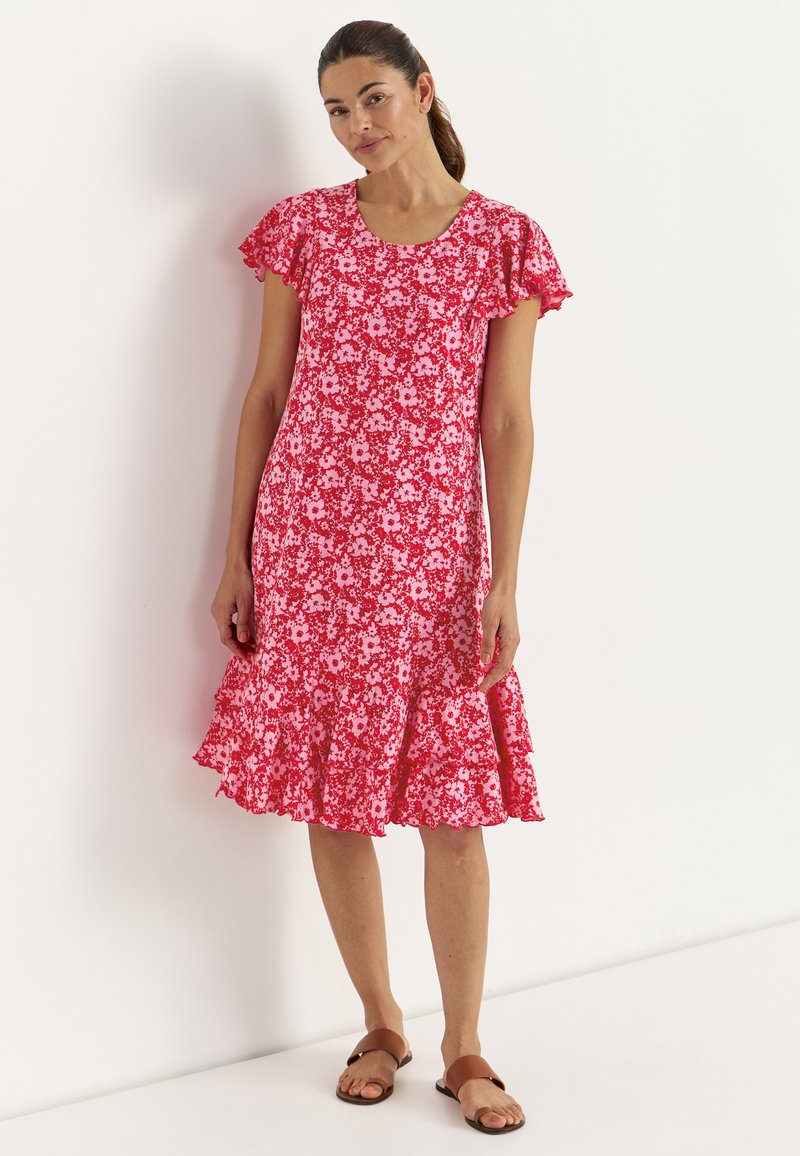 Kvinde med mørkt hår i hestehale iført en rød og pink blomsterkjole med flæser på ærmer og skørt, stående op ad en hvid væg.