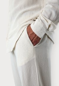 Hellbeige Leinenhemd mit lockerer Passform, langen Ärmeln, durchgehender Knopfleiste und seitlichen Taschen auf passenden Leinenhosen.