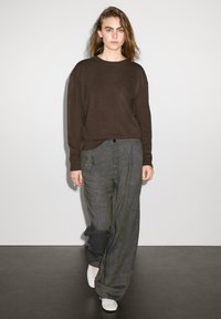 Pull en tricot marron, pantalon gris ample avec poches latérales et baskets blanches. Texture lisse, coupe décontractée et design minimal.