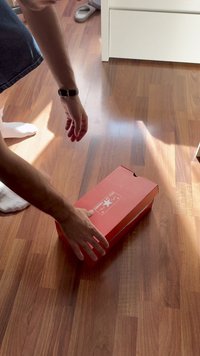 Červená kartónová krabica na topánky na drevenej podlahe s bielou etiketou obsahujúcou logo "Converse All Star". Ruka sa snaží zdvihnúť krabicu.