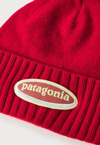 Gorro de lana rojo con textura acanalada. Presenta un parche con un diseño ovalado y la palabra "Patagonia" en costura amarilla.