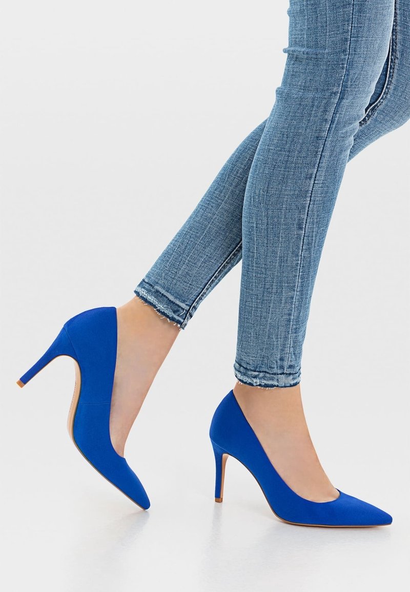 Des escarpins bleus en tissu à talons hauts, avec un bout pointu et une silhouette élégante, associés à un jean en denim bleu clair, coupé au niveau de la cheville.