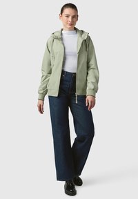 Jasnozielona kurtka zapinana na zamek błyskawiczny z kapturem, z elastycznymi mankietami, noszona na białym golfie. Zestawiona z szerokimi ciemnoniebieskimi dżinsami i czarnymi butami.