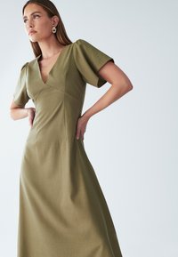 Ženska v olivno zeleni obleki s kratkimi rokavi in V-izrezom, z rokami na bokih, gleda rahlo levo proti enotni svetli ozadju.