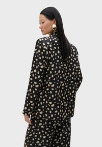 Crni blejzer s bež apstraktnim točkicama, krojena fit, glatka tkanina i izrez na leđima. Uparen s odgovarajućim hlačama.