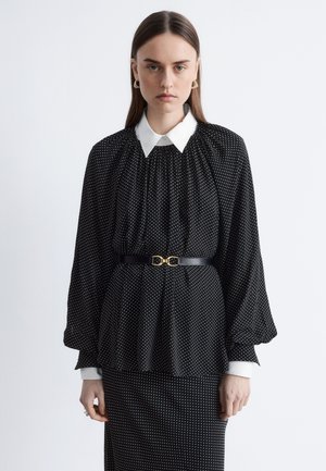Black polka dot blouse with a white collar, featuring puffed sleeves and a fitted waist accentuated by a black belt with a gold buckle.