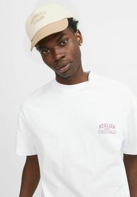 Witte T-shirt met korte mouwen met paarse borduursels van "ATELIER ORIGINALS" op de borst. Crèmekleurige en beige pet met geborduurde tekst.