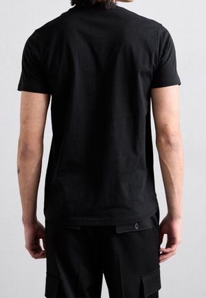 Osoba o średnim odcieniu skóry ubrana w gładką, czarną koszulkę z krótkim rękawem i czarne spodnie, widziana od tyłu na jednolitym tle.