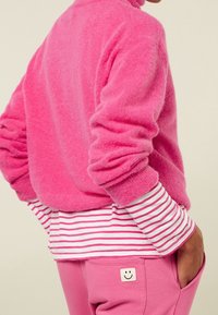Maglione rosa soffice con maniche arrotolate sopra una camicia a maniche lunghe bianca e rosa a righe, che completa dei pantaloni rosa con un sorriso ricamato.